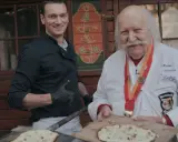 Deux chefs, l'un jeune et souriant, l'autre âgé avec une médaille, présentent des tartes flambées, évoquant l'origine du flamenkuch.