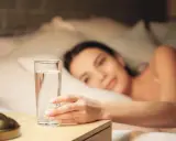Une main attrape un verre d'eau posé à droite sur une table de chevet, une femme se réveille en arrière-plan.