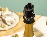 Un bouchon noir sur une bouteille de champagne dorée, prêt à servir. Des huîtres et des verres de champagne en arrière-plan.