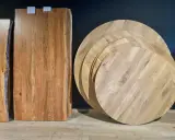 Plusieurs plateaux de table ronde en bois, empilés, illustrent la définition d'une table ronde. Des planches de bois brut sont aussi visibles.