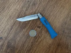 Un couteau Opinel bleu personnalisé "Marie" est posé à côté d'une pièce de 2 euros. Un cadeau symbolique, comme offrir un couteau pour donner une pièce.