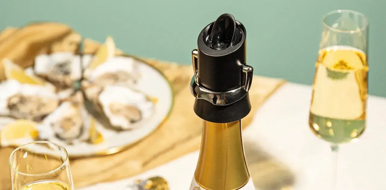Un bouchon noir sur une bouteille de champagne dorée, prêt à servir. Des huîtres et des verres de champagne en arrière-plan.
