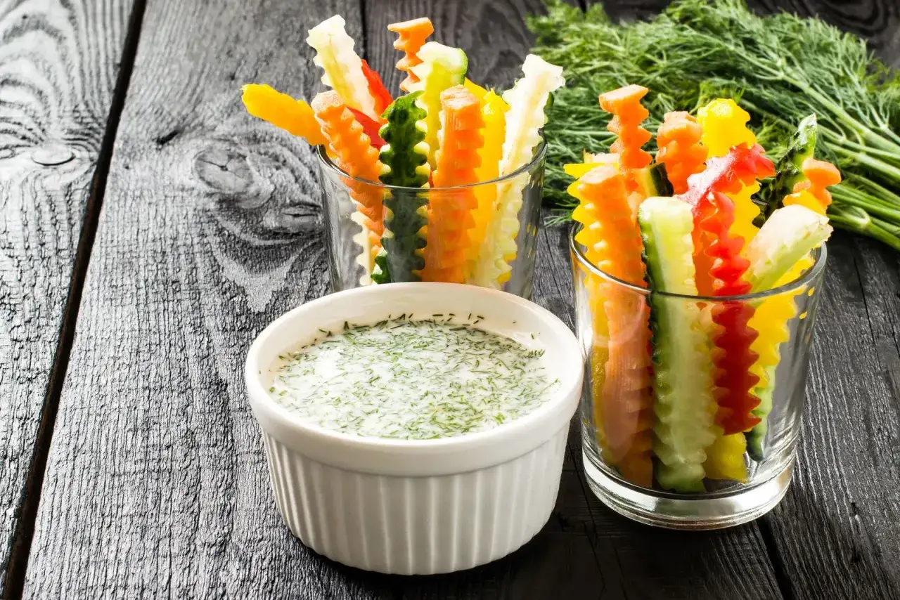 Légumes coupés en bâtonnets colorés dans des verres, accompagnés d'une sauce crémeuse aux herbes. Une idée de dips recette facile et saine.