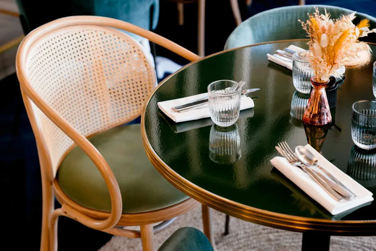 Une table de bistrot avec des couverts et un verre d'eau, à côté d'une chaise en rotin, évoquant une ancienne tradition française.