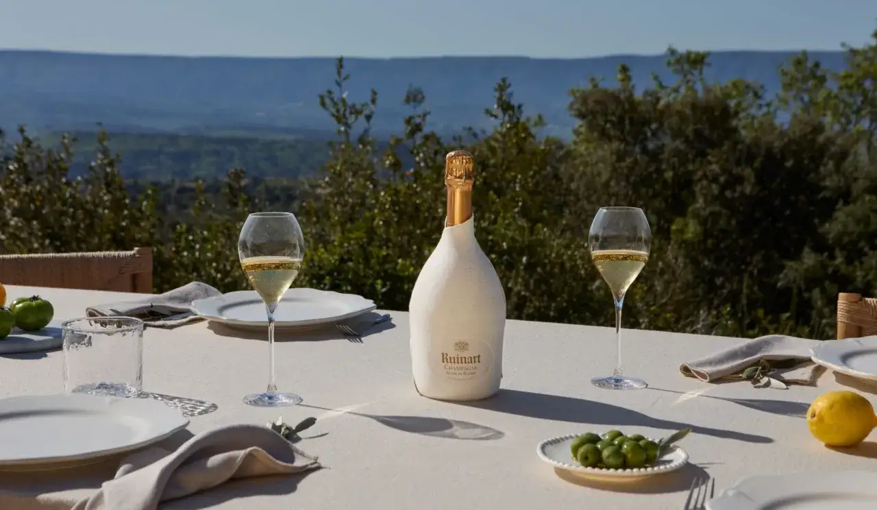 Une bouteille de champagne Ruinart sur une table dressée en extérieur, avec deux verres remplis. La question