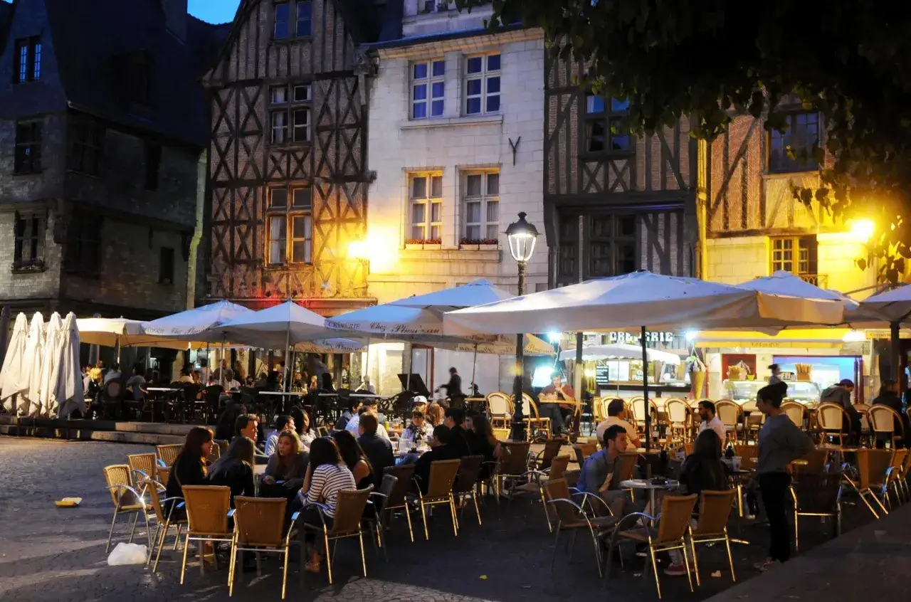 Ambiance animée d'un restaurant sur Tours, avec des tables dressées sous des parasols, des gens attablés et des bâtiments anciens illuminés.