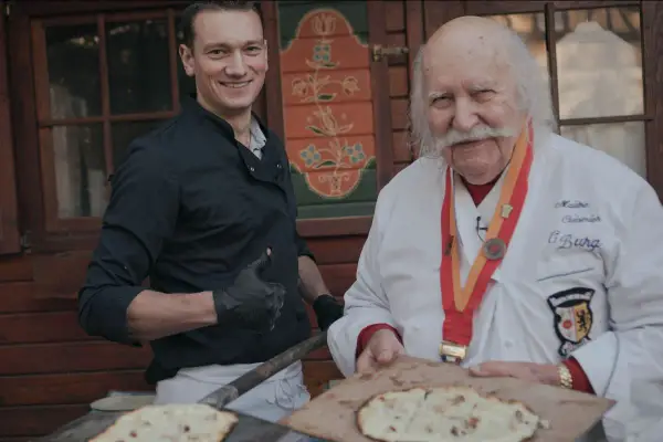 Deux chefs, l'un jeune et souriant, l'autre âgé avec une médaille, présentent des tartes flambées, évoquant l'origine du flamenkuch.
