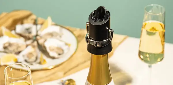 Un bouchon noir sur une bouteille de champagne dorée, prêt à servir. Des huîtres et des verres de champagne en arrière-plan.