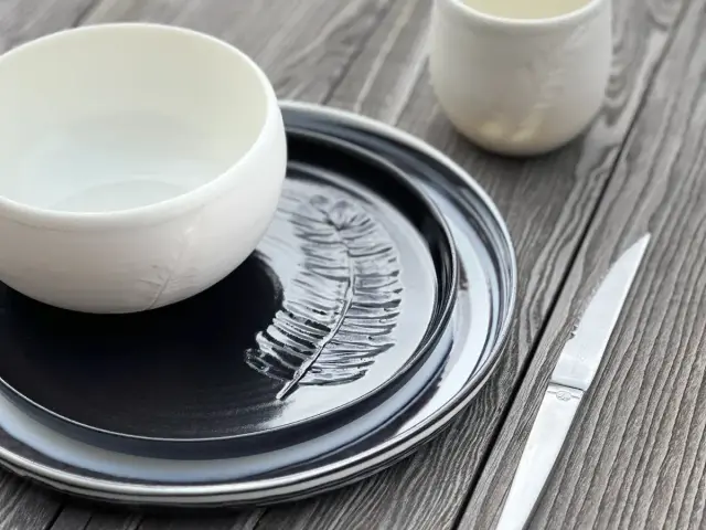 Service de table en porcelaine ou céramique, avec une assiette noire gravée d'une plume, un bol blanc et une tasse assortie.