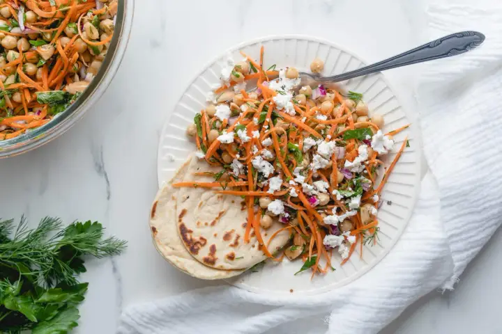 Salade de pois chiches et carottes râpées assaisonnement frais, garnie de feta émiettée et servie avec du pain plat.
